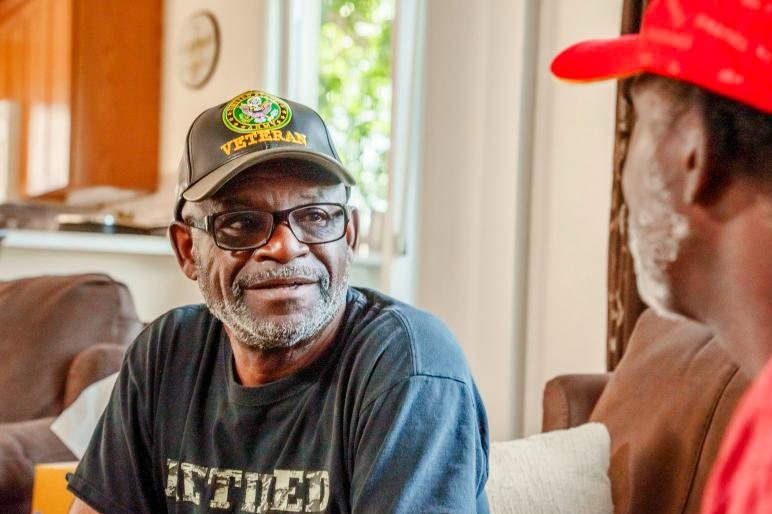 A smiling Army veteran in a veteran cap talking with a friend